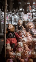 A collection of dolls is on display, some wrapped in plastic. The dolls have various hair colors and styles, and they are positioned on a street vendor's stall. In the background, there are multiple blurred objects and lights.