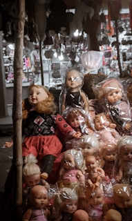 A collection of dolls is on display, some wrapped in plastic. The dolls have various hair colors and styles, and they are positioned on a street vendor's stall. In the background, there are multiple blurred objects and lights.