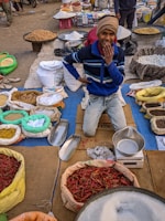 A smiling vendor sits on a mat surrounded by numerous sacks and baskets filled with spices, grains, and other items at a market. The vendor is wearing a blue striped sweater and a beige beanie, with one hand thoughtfully touching his chin. The scene is colorful with various items like red chilies, white grains, and legumes displayed around him.