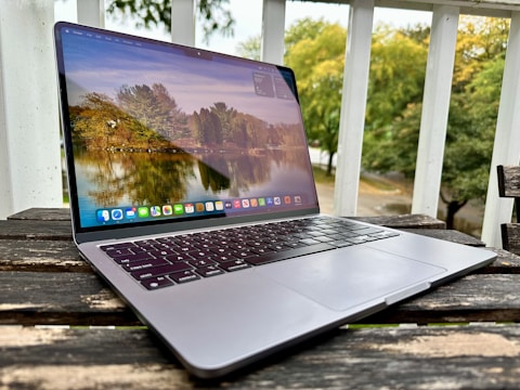 A laptop is placed on a wooden table outdoors, with a scenic nature wallpaper featuring trees and a lake visible on the screen. The setting appears to be a patio or balcony with white railings and greenery in the background.