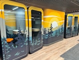 Colorful office pods with nature-themed designs, featuring a sunset over pine trees. The interior includes a small desk and chair, visible through glass doors. The flooring is a combination of wood and carpet.