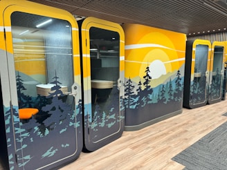 Colorful office pods with nature-themed designs, featuring a sunset over pine trees. The interior includes a small desk and chair, visible through glass doors. The flooring is a combination of wood and carpet.