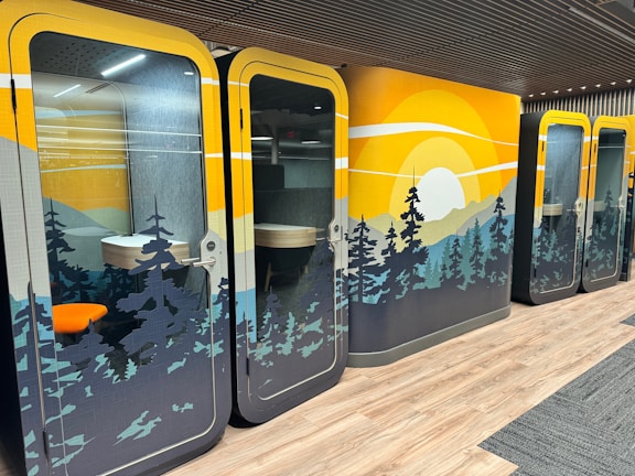 Colorful office pods with nature-themed designs, featuring a sunset over pine trees. The interior includes a small desk and chair, visible through glass doors. The flooring is a combination of wood and carpet.