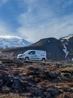 A white van with visible branding is parked on a rocky landscape with sparse vegetation. In the background, a large mountain range is partially covered in snow, with a clear blue sky above.