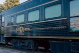 A vintage train car with the name 'Jose Cuervo Express' displayed prominently in decorative gold lettering. The train is dark blue with horizontal lines running along the body, and the windows have a light tint. The setting is outdoors with visible greenery and utility poles in the background.
