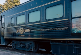 A vintage train car with the name 'Jose Cuervo Express' displayed prominently in decorative gold lettering. The train is dark blue with horizontal lines running along the body, and the windows have a light tint. The setting is outdoors with visible greenery and utility poles in the background.