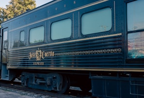 A vintage train car with the name 'Jose Cuervo Express' displayed prominently in decorative gold lettering. The train is dark blue with horizontal lines running along the body, and the windows have a light tint. The setting is outdoors with visible greenery and utility poles in the background.