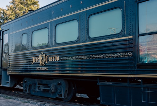 A vintage train car with the name 'Jose Cuervo Express' displayed prominently in decorative gold lettering. The train is dark blue with horizontal lines running along the body, and the windows have a light tint. The setting is outdoors with visible greenery and utility poles in the background.