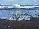 A clear, transparent ice block rests on a dark sandy beach with smooth pebbles scattered around. In the background, turquoise ocean waves gently crash under a partially cloudy sky.