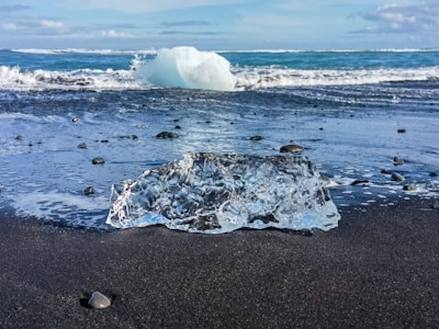 A clear, transparent ice block rests on a dark sandy beach with smooth pebbles scattered around. In the background, turquoise ocean waves gently crash under a partially cloudy sky.