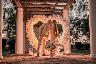A couple is kissing under a heart-shaped floral arch, surrounded by lit candles. They are elegantly dressed in light-toned outfits, and the setting features tall white columns and a lush garden backdrop.