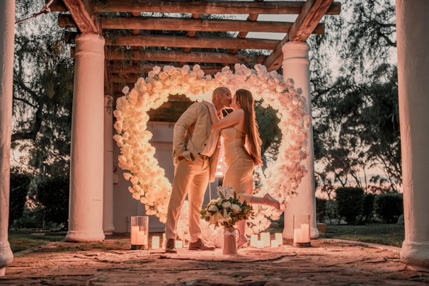 A couple is kissing under a heart-shaped floral arch, surrounded by lit candles. They are elegantly dressed in light-toned outfits, and the setting features tall white columns and a lush garden backdrop.
