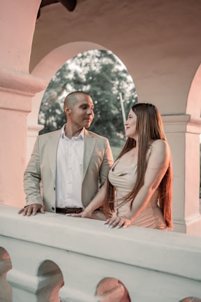 A man and a woman are standing on a balcony with an arched architectural design behind them. They are dressed formally, with the man wearing a light-colored blazer and the woman in a fitted, sleeveless dress. They are gazing at each other with smiles, suggesting a moment of connection or affection between them. The background features soft, muted colors and natural greenery visible through the arches.