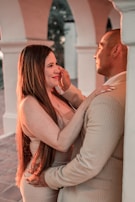 A couple is embracing each other in a warm, intimate setting. The woman, with long hair, has a gentle smile and is touching the man's face. The man, dressed in a light-colored suit, holds the woman close with a tender expression. The background features arches and soft lighting, creating a romantic atmosphere.