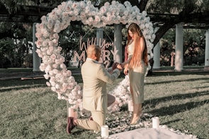 A romantic outdoor marriage proposal with a man kneeling and presenting a ring to a woman. They are surrounded by an ornate floral heart arrangement, with words 'Will You Marry Me?' illuminated behind. The setting includes green grass and white columns, suggesting a garden or park.