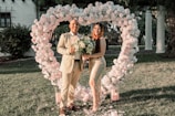 A couple stands in front of a large heart-shaped floral arrangement made of white and pink roses in an outdoor setting. They are dressed in formal attire, with the man in a beige suit and the woman in an elegant dress. The woman holds a bouquet of flowers while the man holds a sign with the words 'She said yes.' The ground is covered in petals, and there are candle holders scattered around.