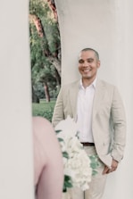 A person in a light-colored suit is smiling while leaning against a light wall. A bouquet of white and green flowers is blurred in the foreground, suggesting a wedding or event setting. Trees with green foliage provide a natural background.