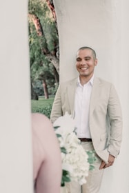 A person in a light-colored suit is smiling while leaning against a light wall. A bouquet of white and green flowers is blurred in the foreground, suggesting a wedding or event setting. Trees with green foliage provide a natural background.