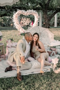 A couple is seated on a picnic setup, with the man lovingly kissing the woman's cheek. They are surrounded by romantic decor including candles, flowers, and a sign that reads, 'Will You Marry Me?' in a heart shape made of flowers. The scene is set outdoors, with greenery in the background and a decorative umbrella adding to the romantic atmosphere.