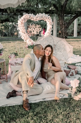A couple is seated on a picnic setup, with the man lovingly kissing the woman's cheek. They are surrounded by romantic decor including candles, flowers, and a sign that reads, 'Will You Marry Me?' in a heart shape made of flowers. The scene is set outdoors, with greenery in the background and a decorative umbrella adding to the romantic atmosphere.