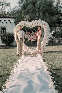 A couple stands at the end of a flower-petal-lined pathway on a grassy lawn. They are framed by a large heart-shaped wreath made of white flowers, with a neon sign in the center asking 'Will you marry me?' The setting suggests a romantic proposal scene with candles placed along the pathway.