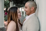 A couple stands closely engaged in conversation, smiling and looking at each other affectionately. They are in an outdoor area with tall white pillars and a covered walkway in the background.