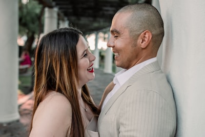 A couple stands closely engaged in conversation, smiling and looking at each other affectionately. They are in an outdoor area with tall white pillars and a covered walkway in the background.