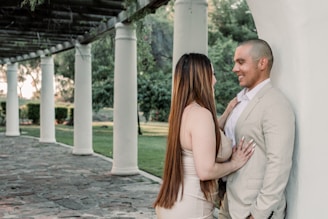 A couple stands under a pergola with white columns and lush greenery in the background. The woman is wearing a light-colored dress, and the man is in a light suit. They appear to be engaged in a conversation, with the woman touching the man's chest.