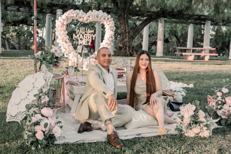 A couple sits on a picnic blanket surrounded by romantic decorations including flowers and a heart-shaped arch with the words 'Will You Marry Me?'. They are dressed elegantly in a park setting with trees and stone pillars in the background.
