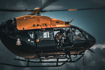 A black and orange helicopter in flight with a soldier or paramilitary personnel inside, wearing a helmet and gear. The helicopter has military insignia on its side and the sky is cloudy and overcast, suggesting an intense or serious atmosphere.