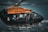 A black and orange helicopter in flight with a soldier or paramilitary personnel inside, wearing a helmet and gear. The helicopter has military insignia on its side and the sky is cloudy and overcast, suggesting an intense or serious atmosphere.