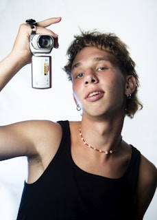 A person with a relaxed expression holds a silver camcorder up to their head. They are wearing a black tank top and a beaded necklace. The background is a simple light color, and the overall lighting is bright.