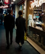 A narrow, dimly lit street with two people walking side by side. They are passing by a shop displaying various goods, and there are neon signs in Arabic above them. The shop window is filled with bags of grains and a salesperson inside is behind a counter.