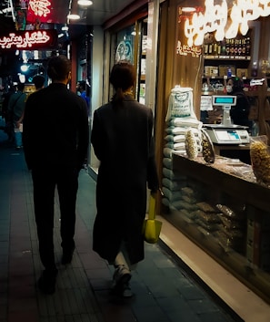A narrow, dimly lit street with two people walking side by side. They are passing by a shop displaying various goods, and there are neon signs in Arabic above them. The shop window is filled with bags of grains and a salesperson inside is behind a counter.