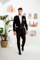 Model wearing a tailored black suit with subtle gold details, standing against a minimalist white backdrop.