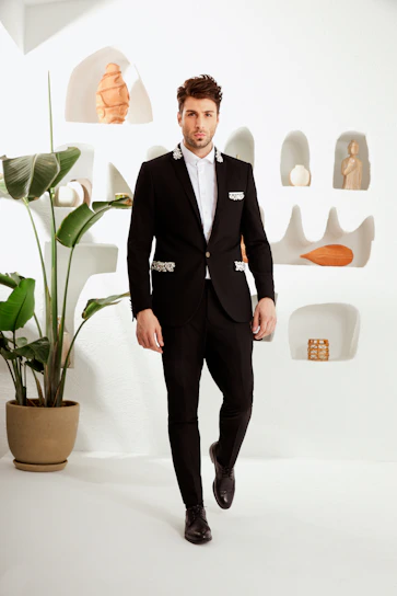 Editorial-style photo of a man wearing a tailored black suit with gold accents, standing against a minimalist beige backdrop.