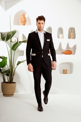 A man in a stylish black suit with decorative white embellishments stands in a minimalist room. The background includes a white wall with recessed niches displaying small sculptures and vases. A large potted plant with broad green leaves adds a touch of nature to the scene.