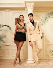 Two people stand elegantly in a luxurious setting with ornate architectural elements. One wears a decorative top and a black fringed skirt, accessorized with large earrings. The other is dressed in a light-colored suit with embellishments. Potted plants and classical columns enhance the opulent atmosphere.