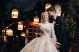 A joyful couple stands together, the woman wearing a glittering wedding dress and headscarf, while the man dons a classic black suit and bow tie. They are smiling warmly at each other, surrounded by softly glowing lanterns that create a romantic atmosphere.