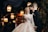 A joyful couple stands together, the woman wearing a glittering wedding dress and headscarf, while the man dons a classic black suit and bow tie. They are smiling warmly at each other, surrounded by softly glowing lanterns that create a romantic atmosphere.