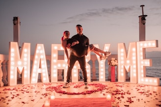 Two people are on a deck by the sea at sunset, surrounded by large illuminated letters spelling 'MARRY ME'. One person is being carried by the other, surrounded by red rose petals on the wooden floor and candles. A red vase with white flowers is placed next to the letters.