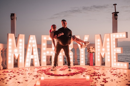 Two people are on a deck by the sea at sunset, surrounded by large illuminated letters spelling 'MARRY ME'. One person is being carried by the other, surrounded by red rose petals on the wooden floor and candles. A red vase with white flowers is placed next to the letters.
