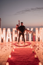 A romantic scene set during sunset with large illuminated letters spelling 'Marry Me' in the background. A man is carrying a woman in his arms, both appearing joyful. The floor is decorated with scattered rose petals and there is a red carpet leading towards the couple, lined with lit candles. There is a bouquet of flowers placed beside the illuminated letters.
