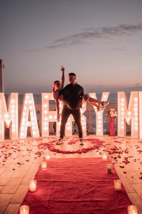 A romantic scene set during sunset with large illuminated letters spelling 'Marry Me' in the background. A man is carrying a woman in his arms, both appearing joyful. The floor is decorated with scattered rose petals and there is a red carpet leading towards the couple, lined with lit candles. There is a bouquet of flowers placed beside the illuminated letters.
