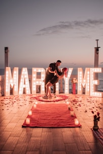 A romantic proposal setting by the sea during sunset. Large illuminated letters spelling 'Marry Me' create a backdrop, surrounded by scattered rose petals and candles on a wooden floor. A couple is captured in a dance pose, adding to the intimate atmosphere.