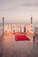 Large illuminated letters spell out 'Marry Me' on a wooden deck overlooking the ocean at sunset. A red carpet leads up to the letters, with candles and rose petals scattered along its path. The scene is framed by white railings and two lantern posts, contributing to a romantic and intimate atmosphere.