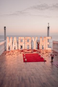 Large illuminated letters spell out 'Marry Me' on a wooden deck overlooking the ocean at sunset. A red carpet leads up to the letters, with candles and rose petals scattered along its path. The scene is framed by white railings and two lantern posts, contributing to a romantic and intimate atmosphere.