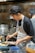 A chef wearing a black shirt and an apron is carefully plating food in a professional kitchen. The background shows various kitchen utensils, pots, and containers. He is focused on arranging the dish with precision, showcasing his culinary skills.