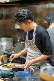 A chef wearing a black shirt and an apron is carefully plating food in a professional kitchen. The background shows various kitchen utensils, pots, and containers. He is focused on arranging the dish with precision, showcasing his culinary skills.