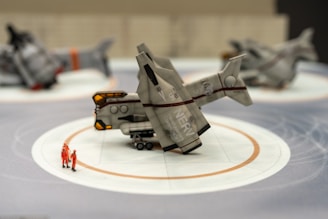 A futuristic spacecraft model is placed on a display stand with two miniature figures in orange suits nearby. The background features two additional spacecraft models, blurred to emphasize the one in the foreground.